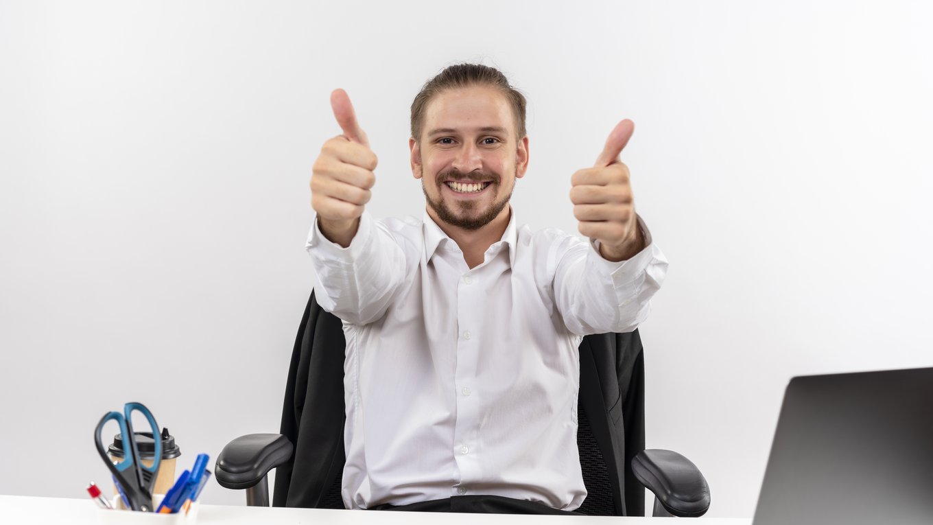 krasivyi-biznesmen-v-beloi-rubaske-glada-v-kameru-ulybaas-pokazyvaet-palec-vverh-sida-za-stolom-v-ofise-na-belom-fone