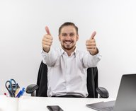 krasivyi-biznesmen-v-beloi-rubaske-glada-v-kameru-ulybaas-pokazyvaet-palec-vverh-sida-za-stolom-v-ofise-na-belom-fone