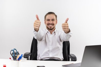 krasivyi-biznesmen-v-beloi-rubaske-glada-v-kameru-ulybaas-pokazyvaet-palec-vverh-sida-za-stolom-v-ofise-na-belom-fone